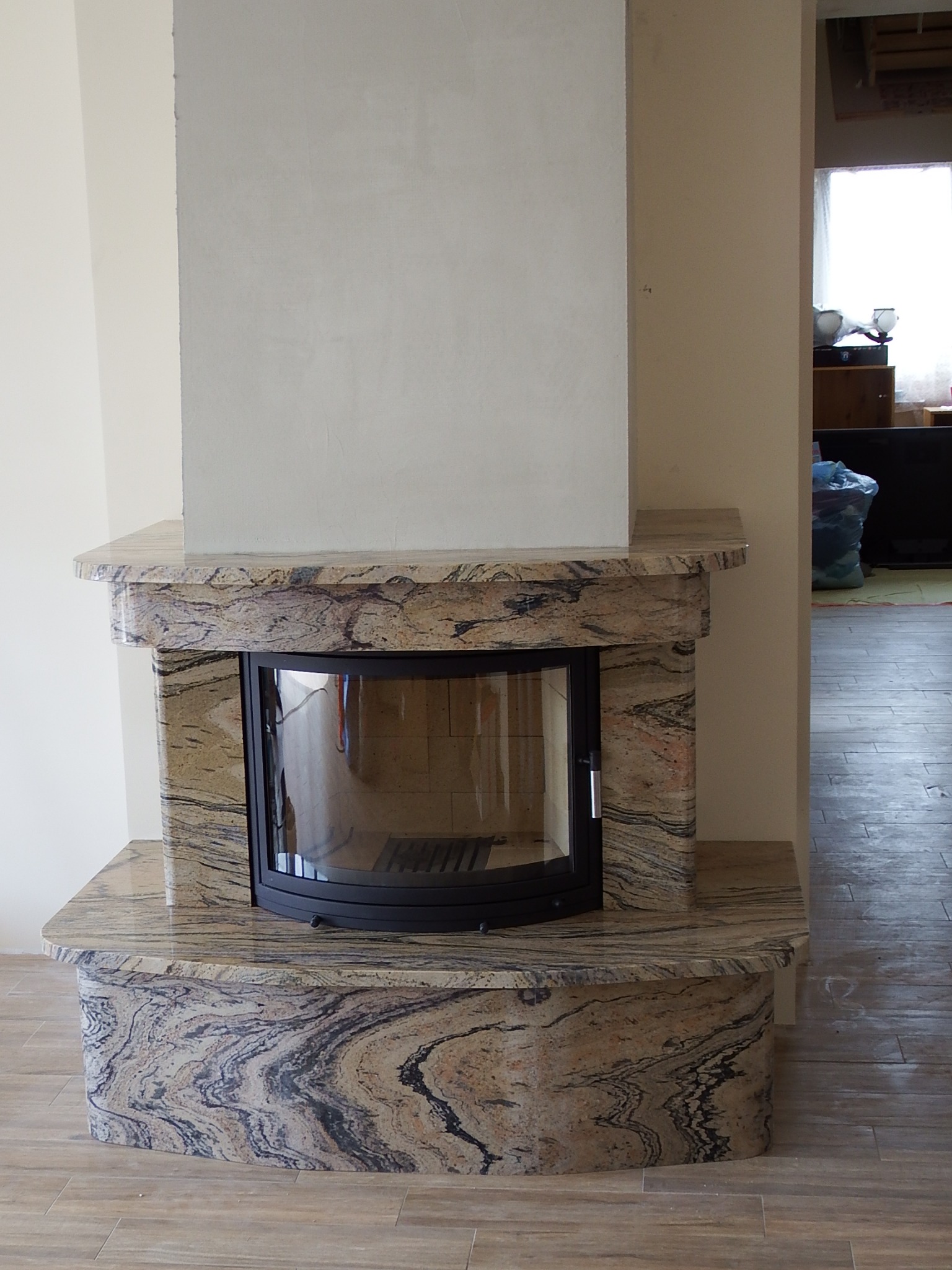 Otynkowany kominek z obudową z kamienia o wyraźnym, falistym wzorze, czarne, półokrągłe drzwiczki, podłoga wyłożona płytkami imitującymi drewno.