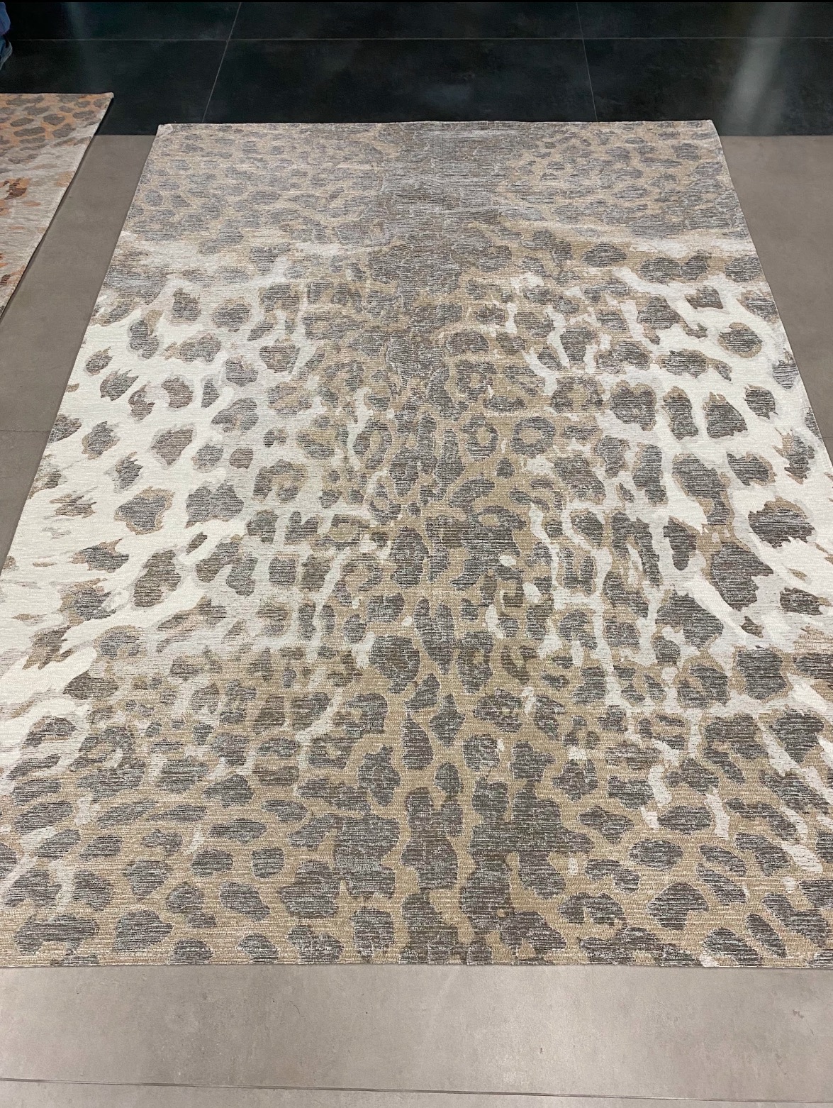 Beżowo-biały dywan z wzorem imitującym panterkę, leżący na szarej podłodze, z czarnym tłem w górnej części kadru.
