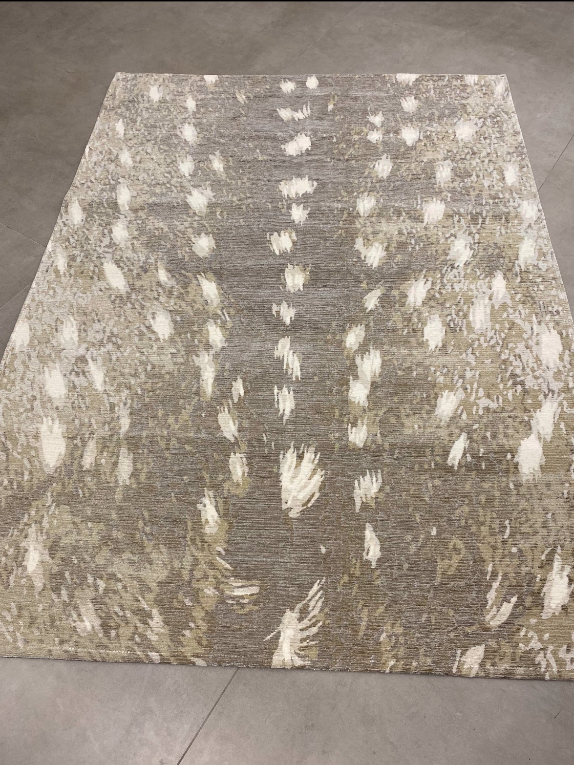 Dywan w odcieniach szarości i beżu z abstrakcyjnym wzorem przypominającym ślady zwierząt, leżący na szarej, betonowej podłodze.