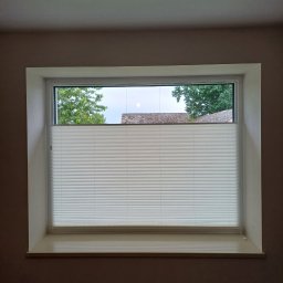 L7 DRZWI - Okno z zamontowaną plisą okienną w kolorze białym, widok na zewnątrz na zielone drzewa i dach budynku.