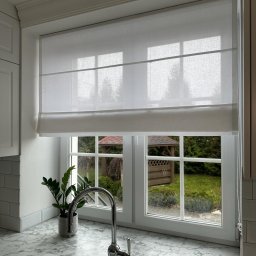 L7 DRZWI - Jasna kuchnia z oknem, na którym zamontowano białą roletę rzymską, widok na ogród z altaną.