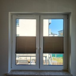 L7 DRZWI - Zamknięte, brązowe żaluzje plisowane w białym oknie PCV z widokiem na balkon i budynki mieszkalne w słoneczny dzień.