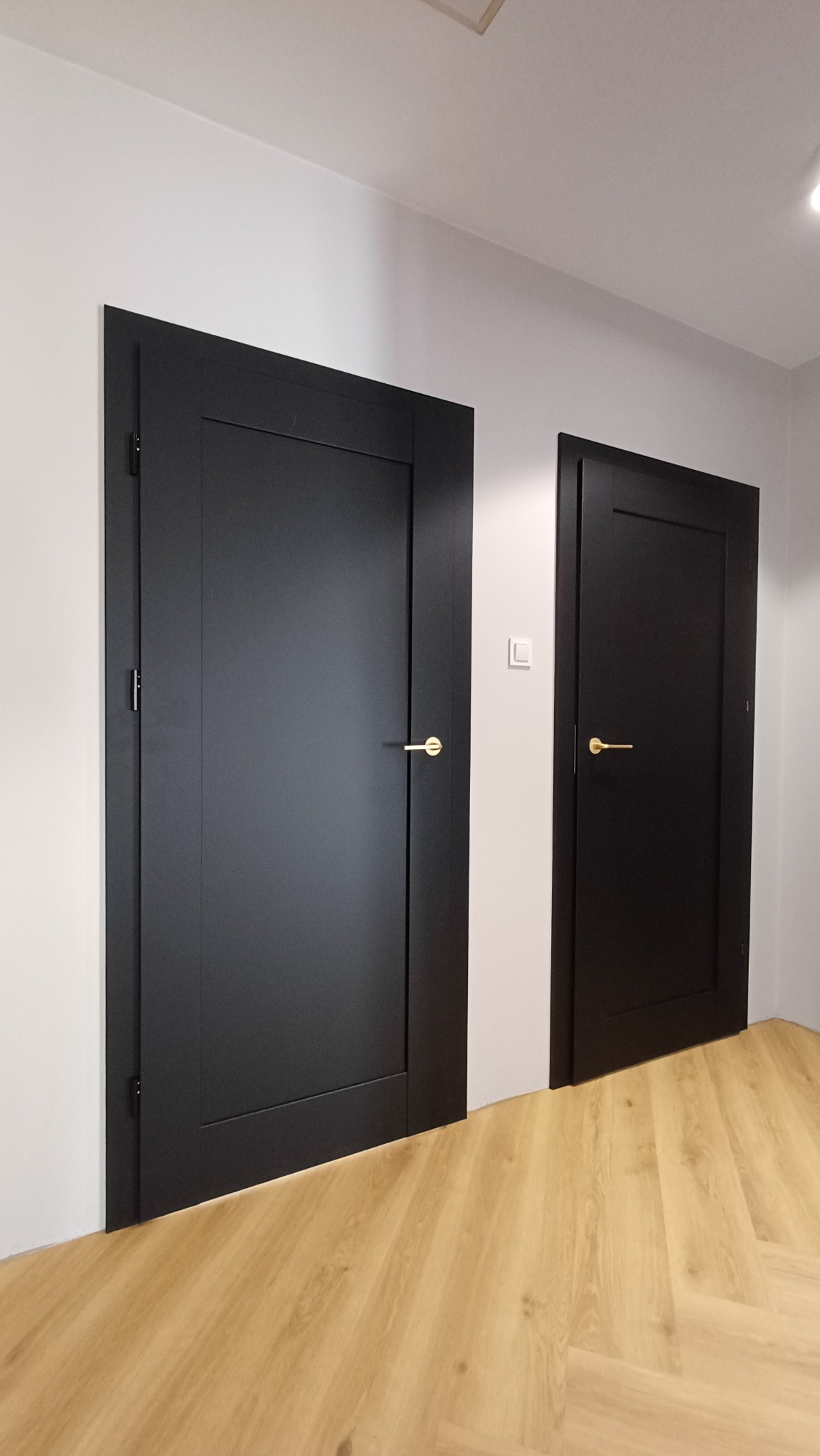 Dwa czarne, frezowane skrzydła drzwiowe z złotymi klamkami, widoczne w korytarzu z podłogą w jodełkę.