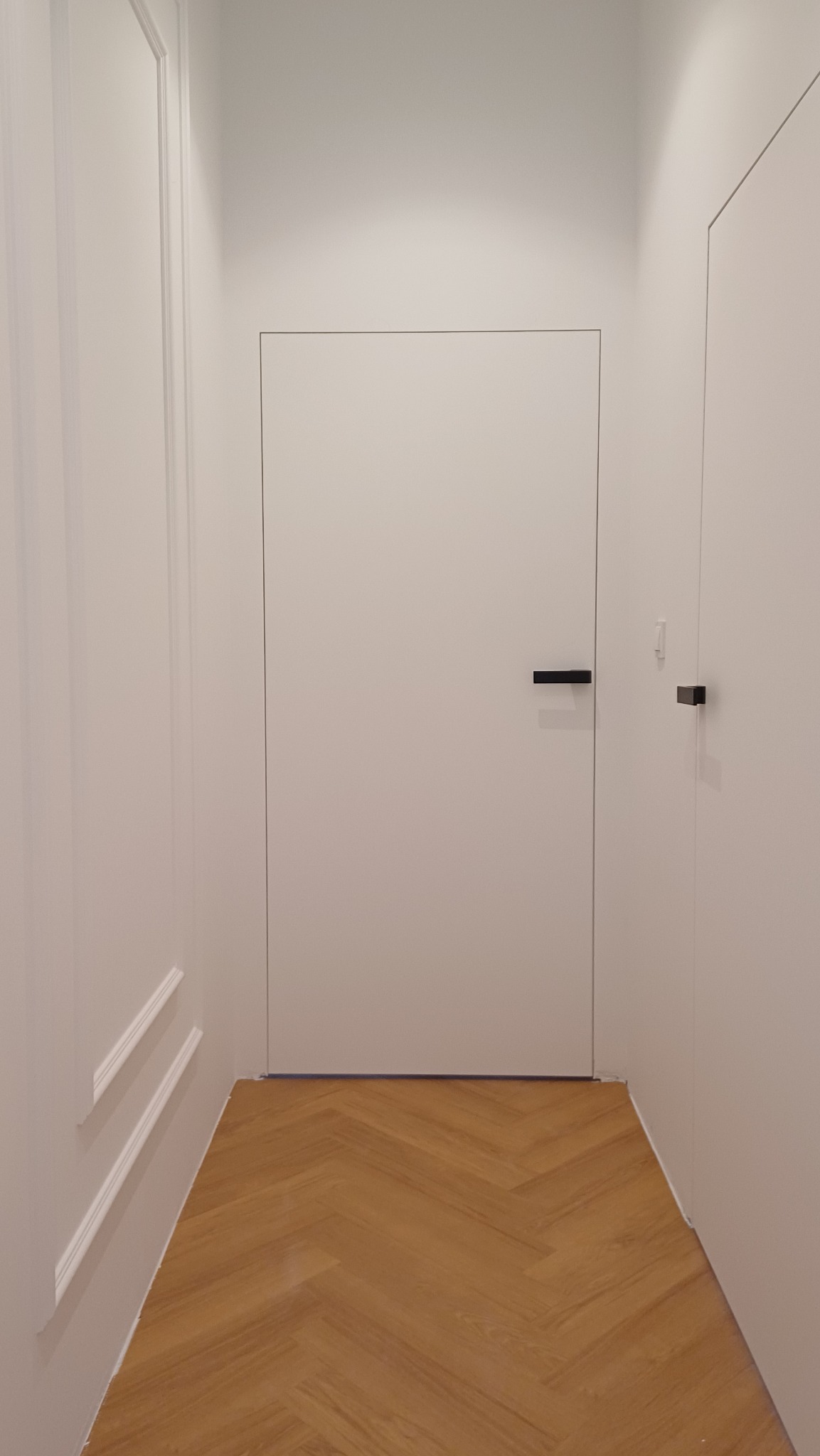 Minimalistyczne, białe skrzydło drzwiowe z ukrytą ościeżnicą i czarną, prostokątną klamką, widoczne w korytarzu z parkietem ułożonym we wzór jodełki.