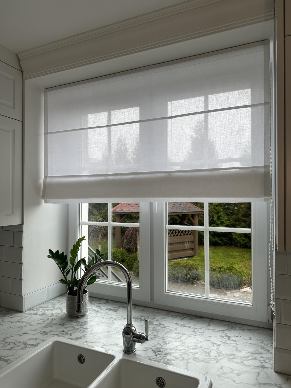 Jasna kuchnia z oknem, na którym zamontowano białą roletę rzymską, widok na ogród z altaną.