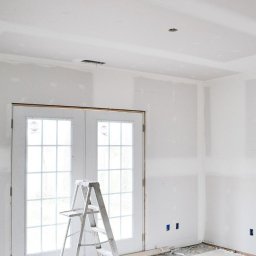 Patronus - Wnętrze w trakcie remontu: ściany pokryte gładzią szpachlową, białe dwuskrzydłowe drzwi balkonowe z szybami, aluminiowa drabina przystawna, widoczne puszki elektryczne w ścianie.