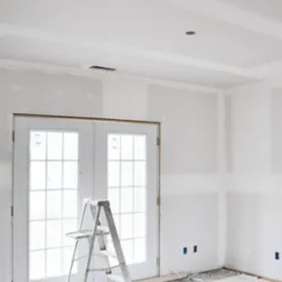 Wnętrze w trakcie remontu: ściany pokryte gładzią szpachlową, białe dwuskrzydłowe drzwi balkonowe z szybami, aluminiowa drabina przystawna, widoczne puszki elektryczne w ścianie.