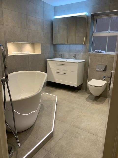 Nowoczesna łazienka z wolnostojącą wanną, szarymi płytkami i podświetleniem LED pod wanną oraz szafką z umywalkami.