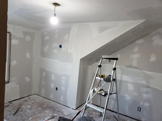 Pokój w trakcie prac wykończeniowych, ściany z nałożoną gładzią szpachlową, widoczna drabina z narzędziami, surowa żarówka zwisająca z sufitu.