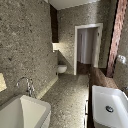 Kamil Malinowski - Nowoczesna łazienka z szarą mozaiką na ścianach i podłodze, białą wanną i umywalką na drewnianym blacie. Widoczne oświetlenie LED i fragment drewnianej belki.