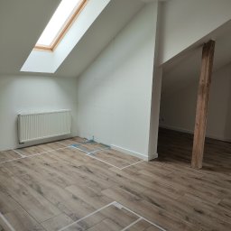 Kamil Malinowski - Podłoga z paneli ułożona w pokoju na poddaszu w Raszynie. Widoczne taśmy wyznaczające obszar roboczy, okno dachowe i grzejnik. Ściany przygotowane pod montaż płyt.