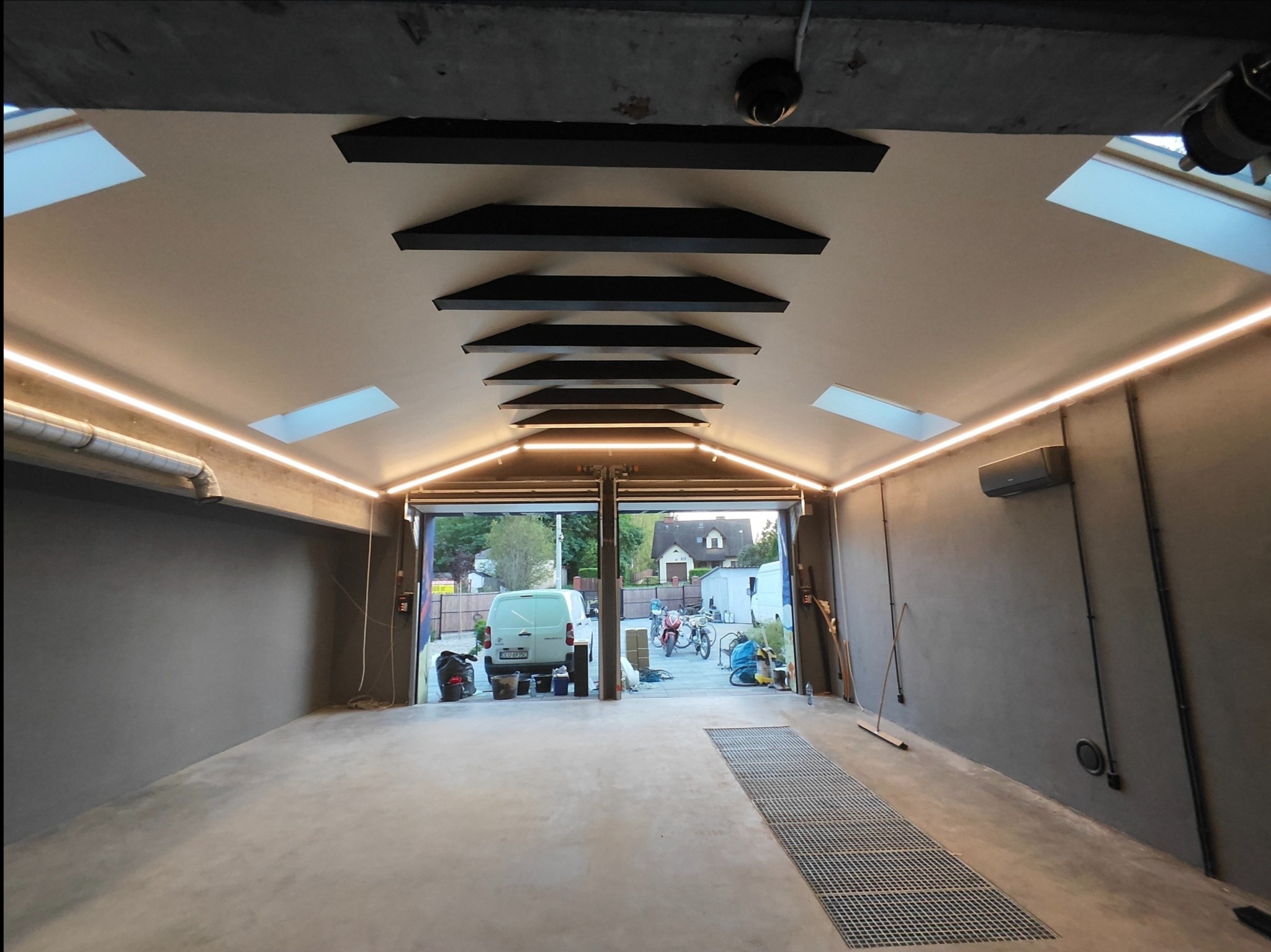 Nowoczesne oświetlenie LED w garażu z szarymi ścianami i betonową podłogą. Widoczne czarne belki na suficie i okna dachowe. Na zewnątrz zaparkowany biały samochód.