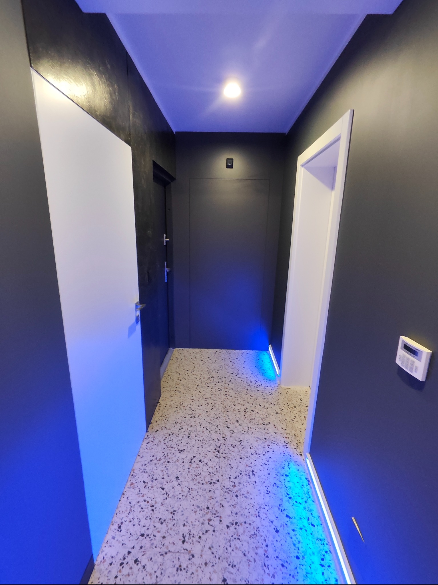 Nowoczesny korytarz po remoncie: ciemne ściany, jasna podłoga lastryko, białe drzwi i listwy LED z niebieskim światłem. Minimalistyczny design.