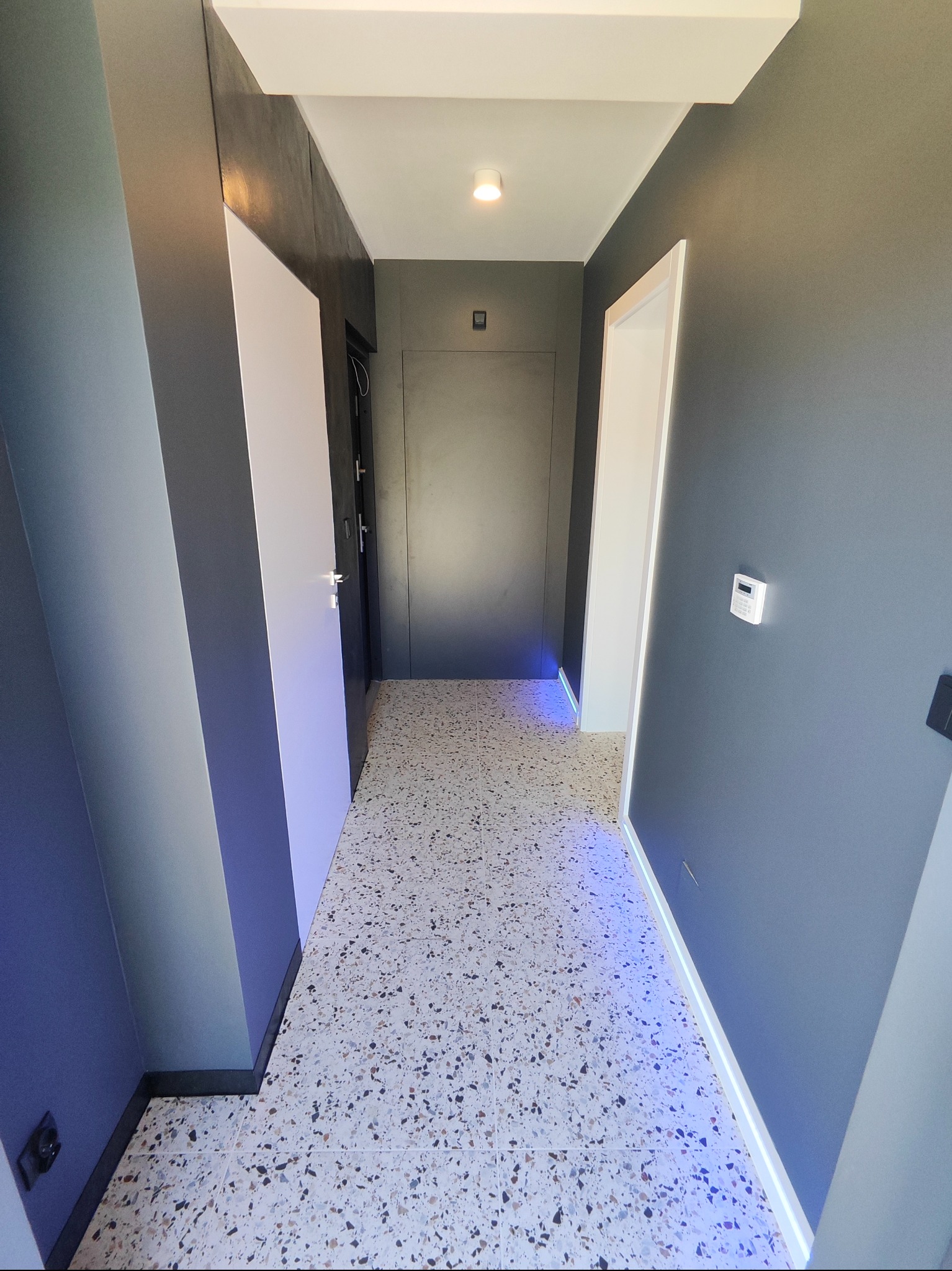 Nowoczesny korytarz po remoncie: szare ściany, terazzo na podłodze, białe drzwi z podświetleniem LED. Minimalistyczny design, czyste linie, widok na drzwi wejściowe.