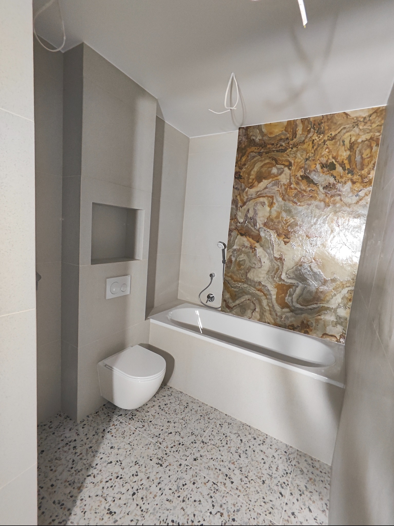 Wykończona łazienka z płytkami imitującymi marmur za wanną, podwieszana toaleta, mozaika na podłodze i wnęka w ścianie. Nowoczesny design.