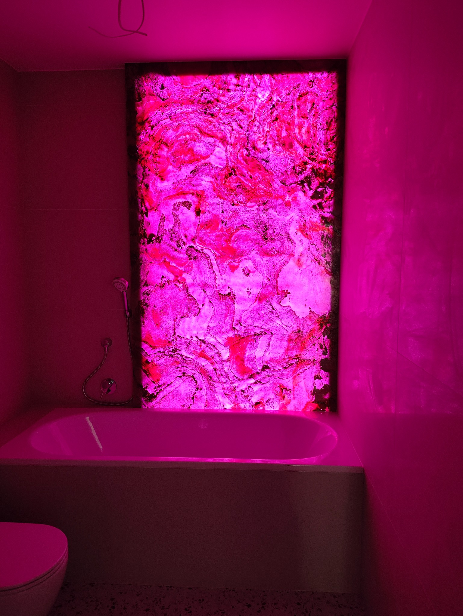 Nowoczesna łazienka z podświetlanym panelem dekoracyjnym w odcieniach różu, wanna i toaleta w tym samym kolorze. Nietypowe oświetlenie tworzy unikalny klimat.