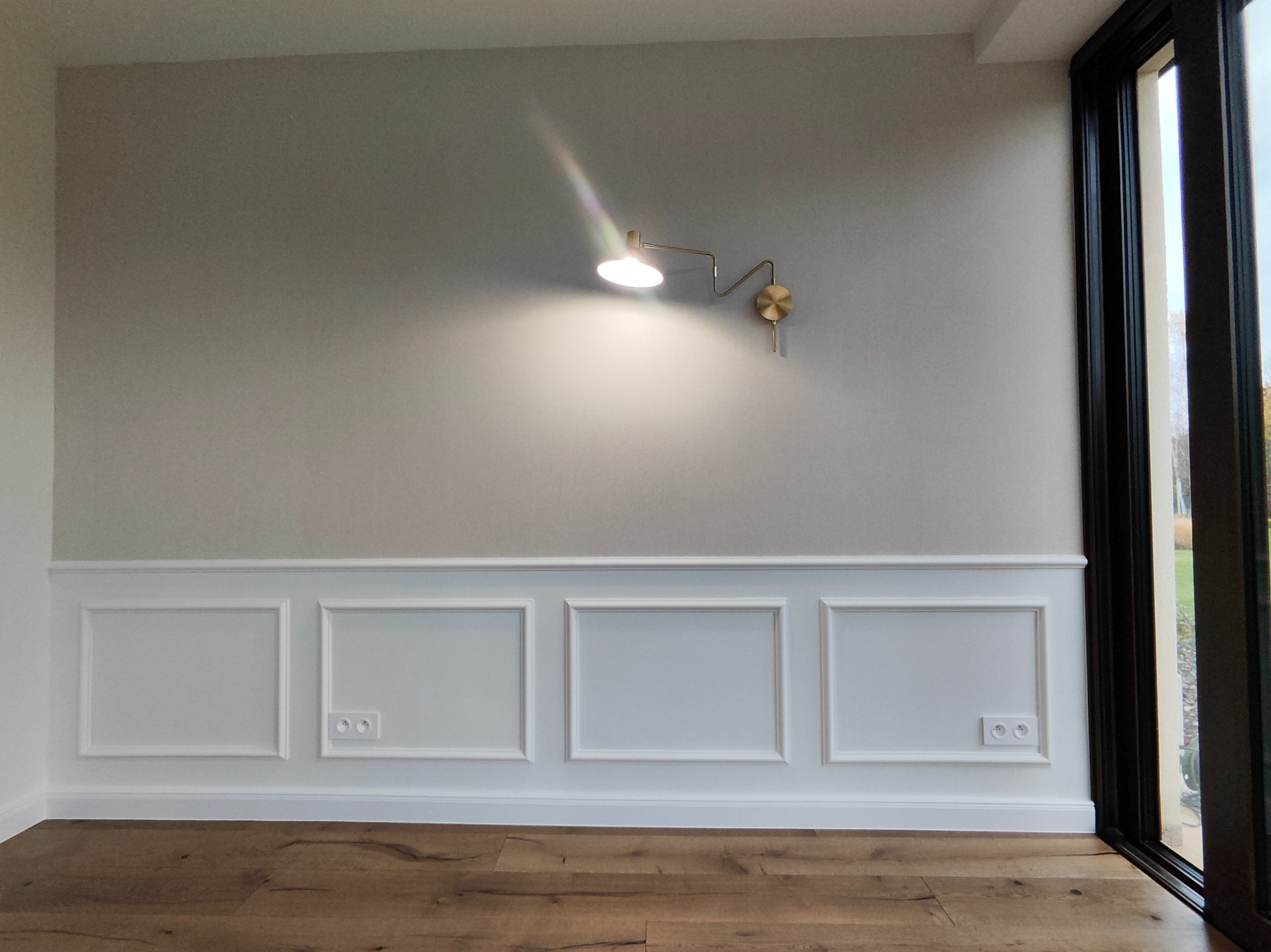 Ściana z dekoracyjną białą boazerią, gładką szarą górą i złotą lampą ścienną. Parkiet na podłodze i czarna rama okienna z prawej strony.