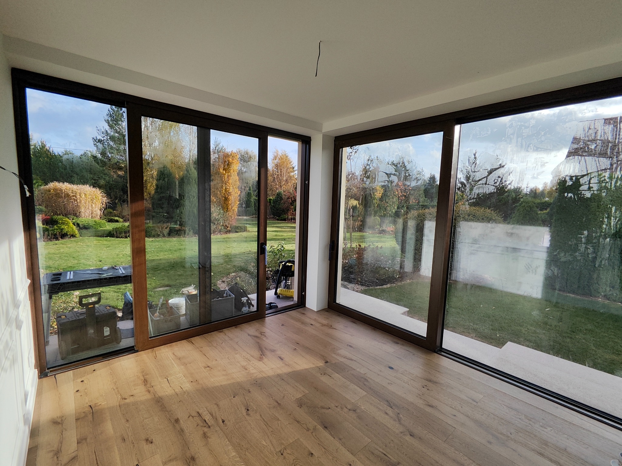 Jasne drewniane panele podłogowe w pokoju z dużymi oknami przesuwnymi, przez które widać zielony ogród. Widoczne narzędzia i akcesoria do montażu.