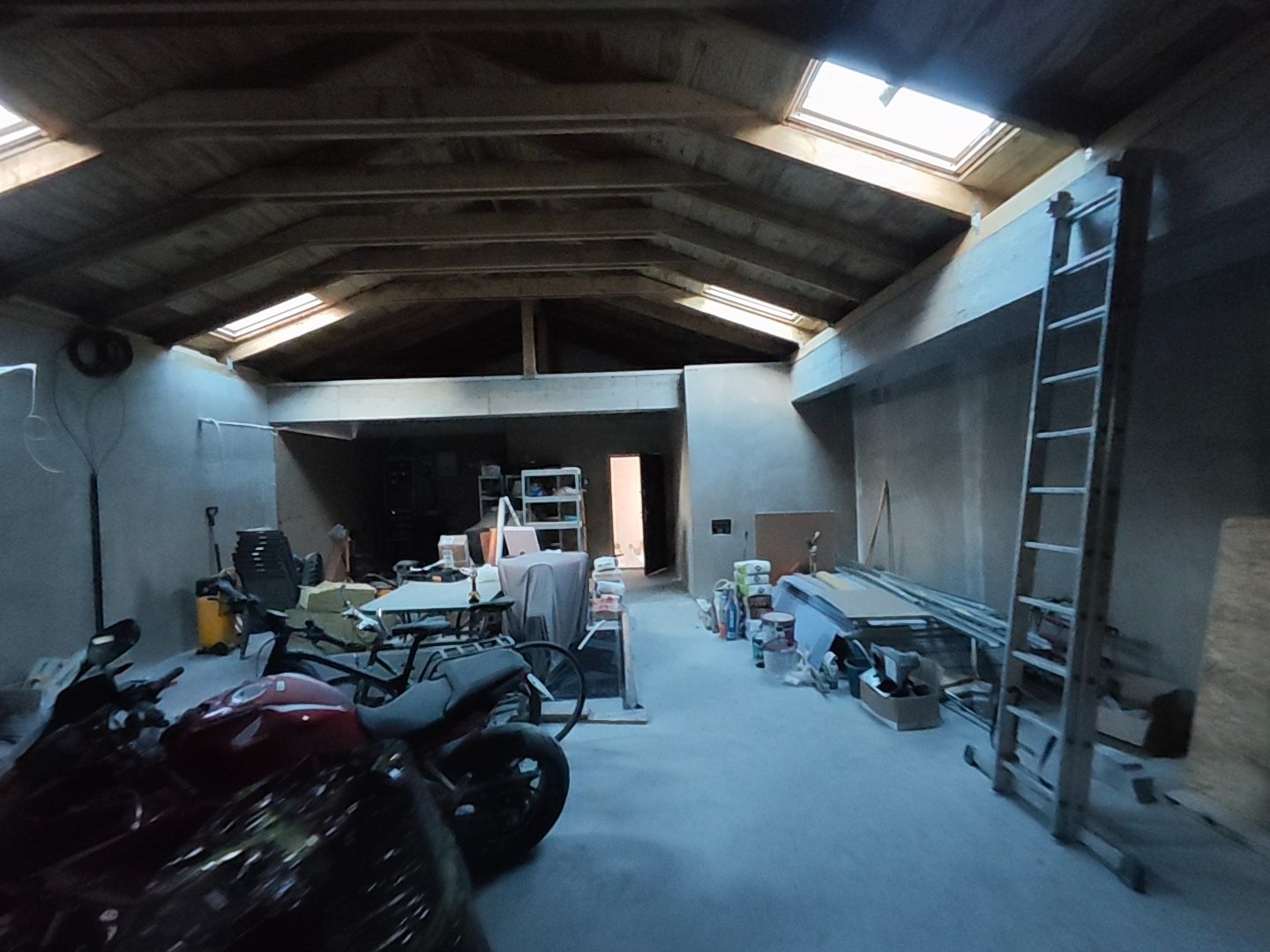 Wnętrze w trakcie prac wykończeniowych: surowy strop z oknami dachowymi, ściany przygotowane do malowania, drabina oparta o ścianę, materiały budowlane i motocykle.
