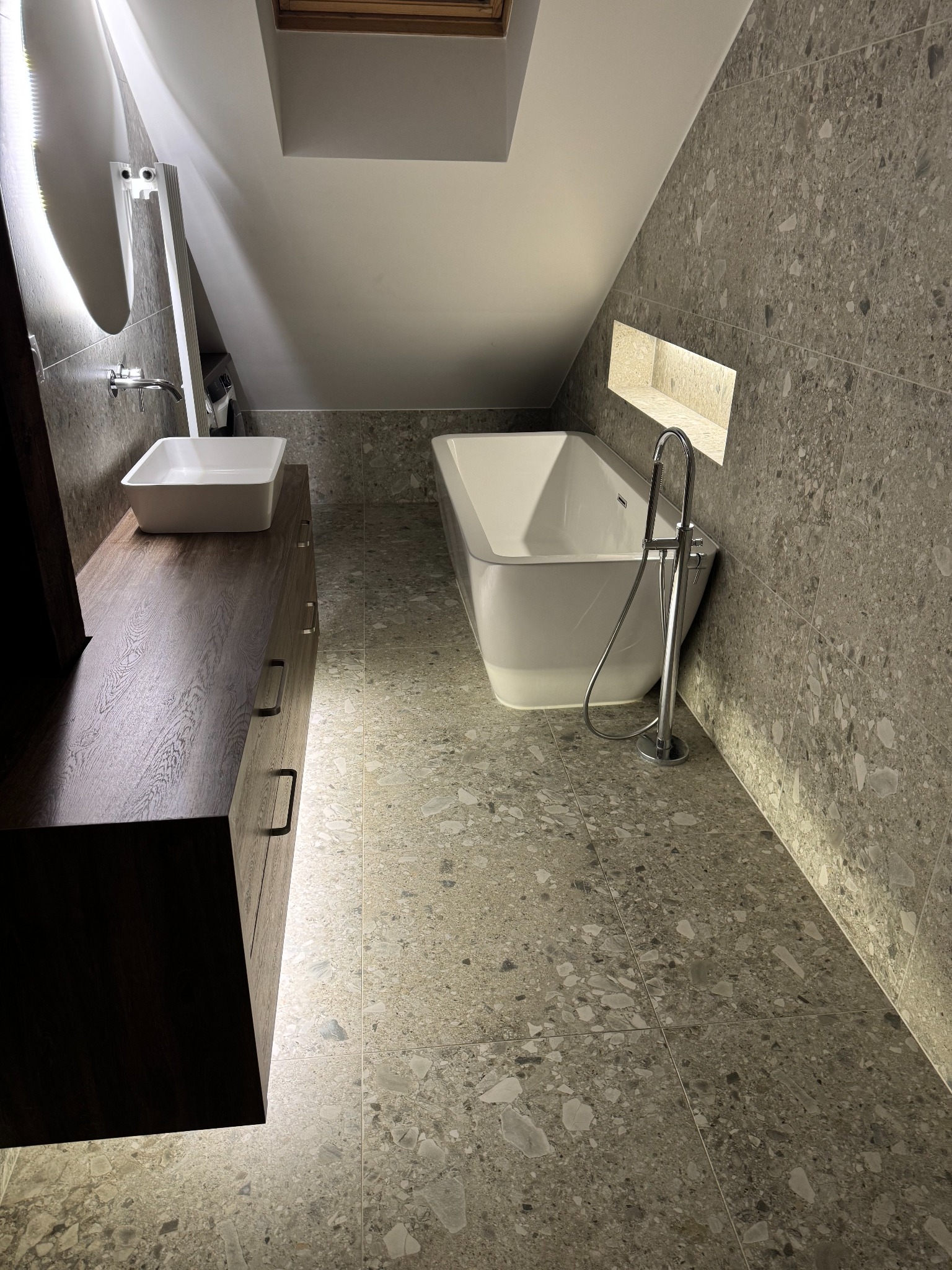 Nowoczesna łazienka na poddaszu z wanną, umywalką na szafce z ciemnego drewna i szarą mozaikową okładziną ścian i podłogi. Oświetlenie LED podkreśla detale.