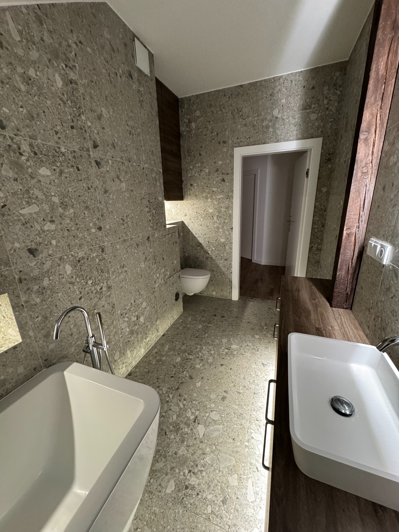 Nowoczesna łazienka z szarą mozaiką na ścianach i podłodze, białą wanną i umywalką na drewnianym blacie. Widoczne oświetlenie LED i fragment drewnianej belki.