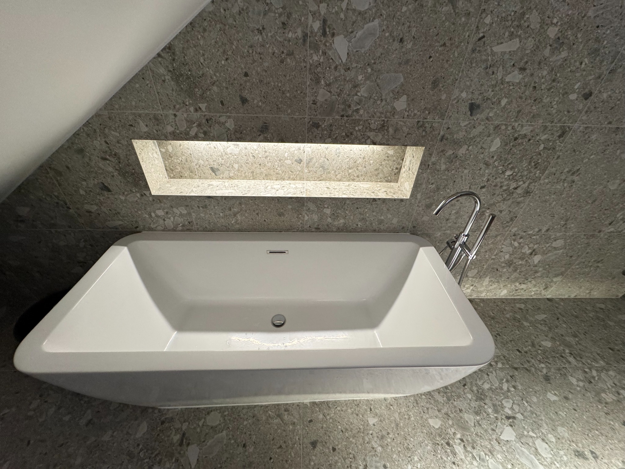 Nowoczesna, biała wanna w łazience z szarymi płytkami lastryko i podświetlaną wnęką. Minimalistyczny design, chromowana bateria. Ukośny sufit dodaje unikalnego charakteru.