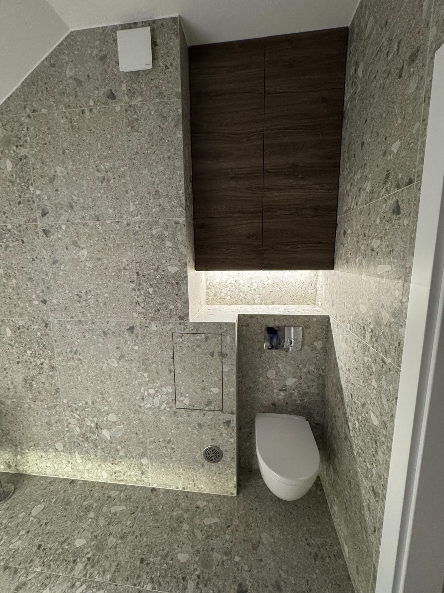 Wykończona łazienka z szarą mozaiką na ścianach i podłodze, podwieszana toaleta, szafka nad toaletą z oświetleniem LED. Nowoczesny design.