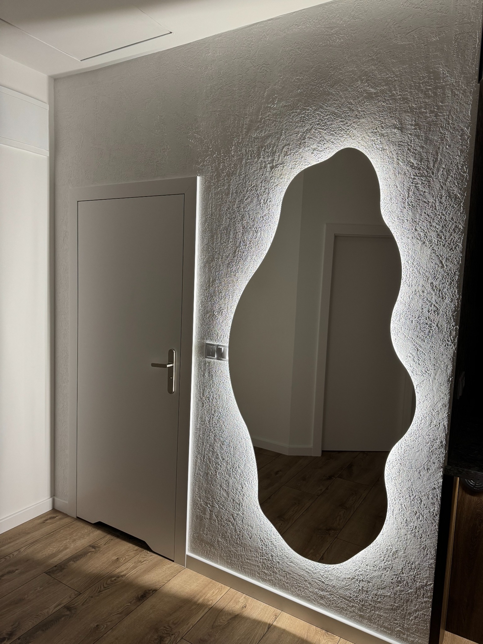 Nowoczesna łazienka: asymetryczne lustro z podświetleniem LED na teksturowanej ścianie, obok drzwi. Parkiet na podłodze. Minimalistyczny design.