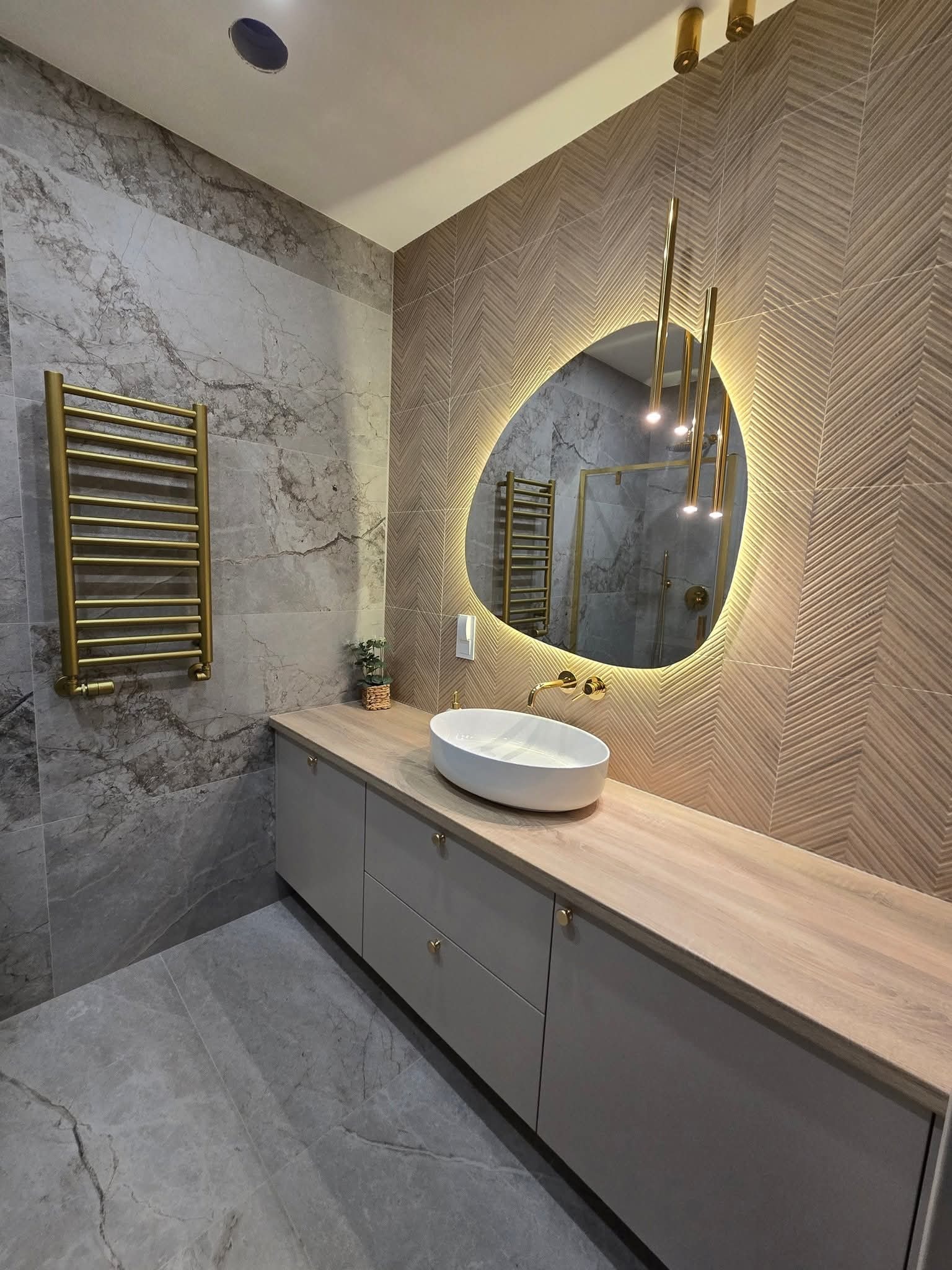 Nowoczesna łazienka z owalnym lustrem z podświetleniem LED, umywalką nablatową i grzejnikiem drabinkowym w kolorze złotym. Płytki imitujące marmur i drewno.