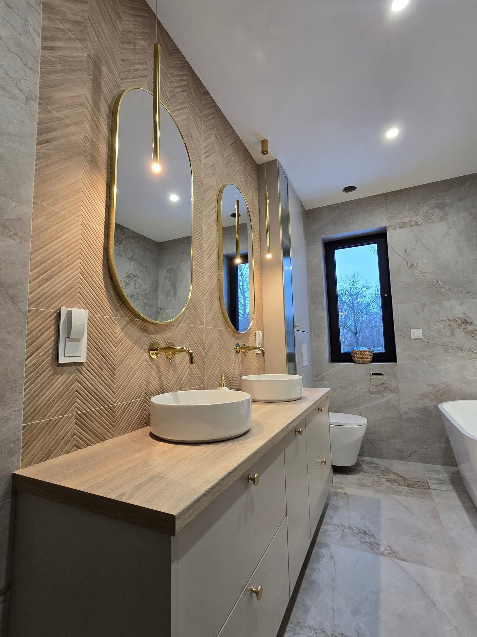 Nowoczesna łazienka z dwoma umywalkami, złotymi detalami i wzorzystą ścianą. Uwagę przykuwają owalne lustra z podświetleniem i marmurowe płytki podłogowe.