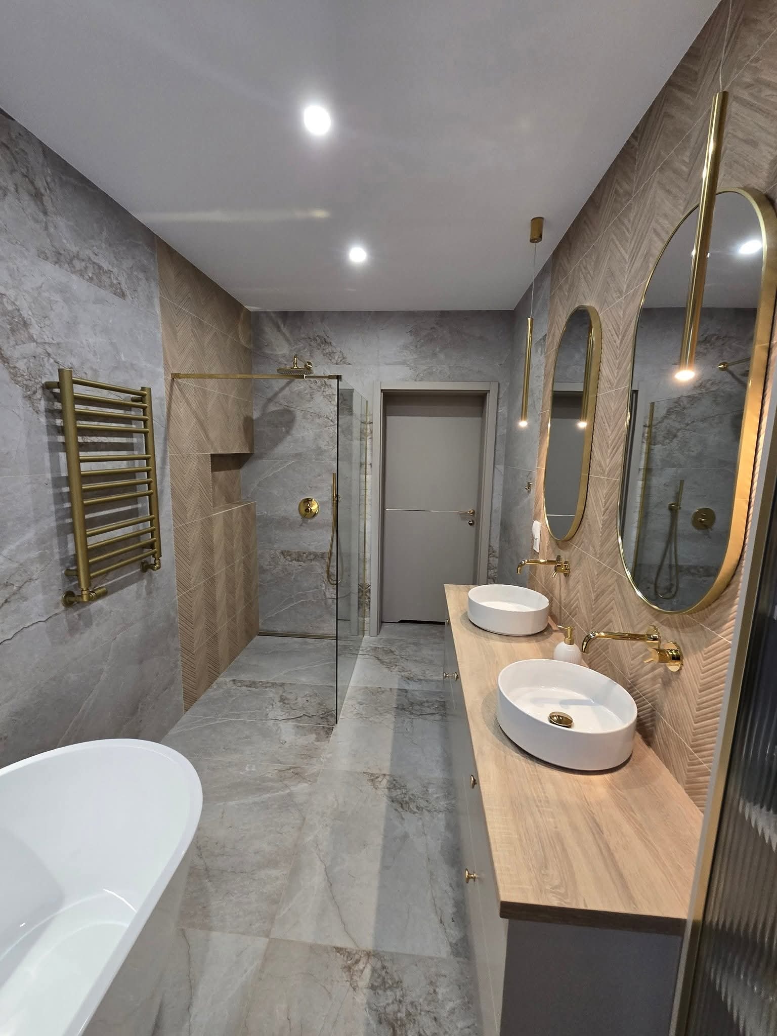 Nowoczesna łazienka z marmurowymi płytkami, złotymi akcentami i owalnymi lustrami. Kabina prysznicowa ze złotą armaturą, wolnostojąca wanna i drewniany blat z umywalkami.