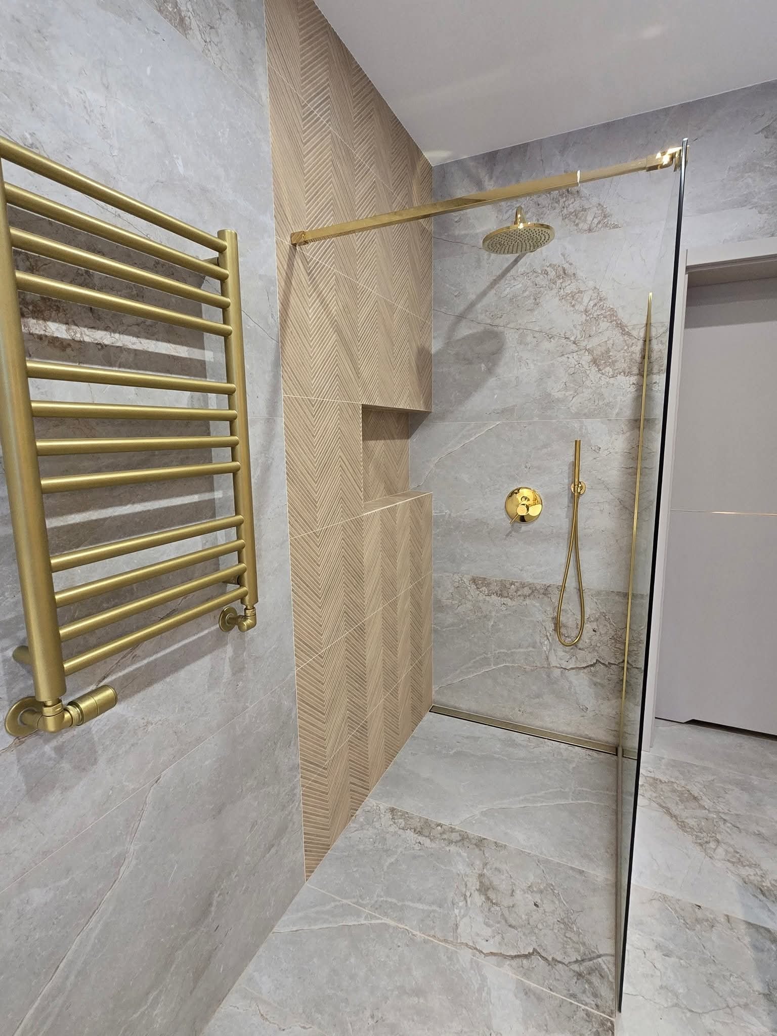 Nowoczesna łazienka z kabiną prysznicową typu walk-in, złota armatura i grzejnik drabinkowy na tle marmurowych płytek. Geometryczny wzór na ścianie.