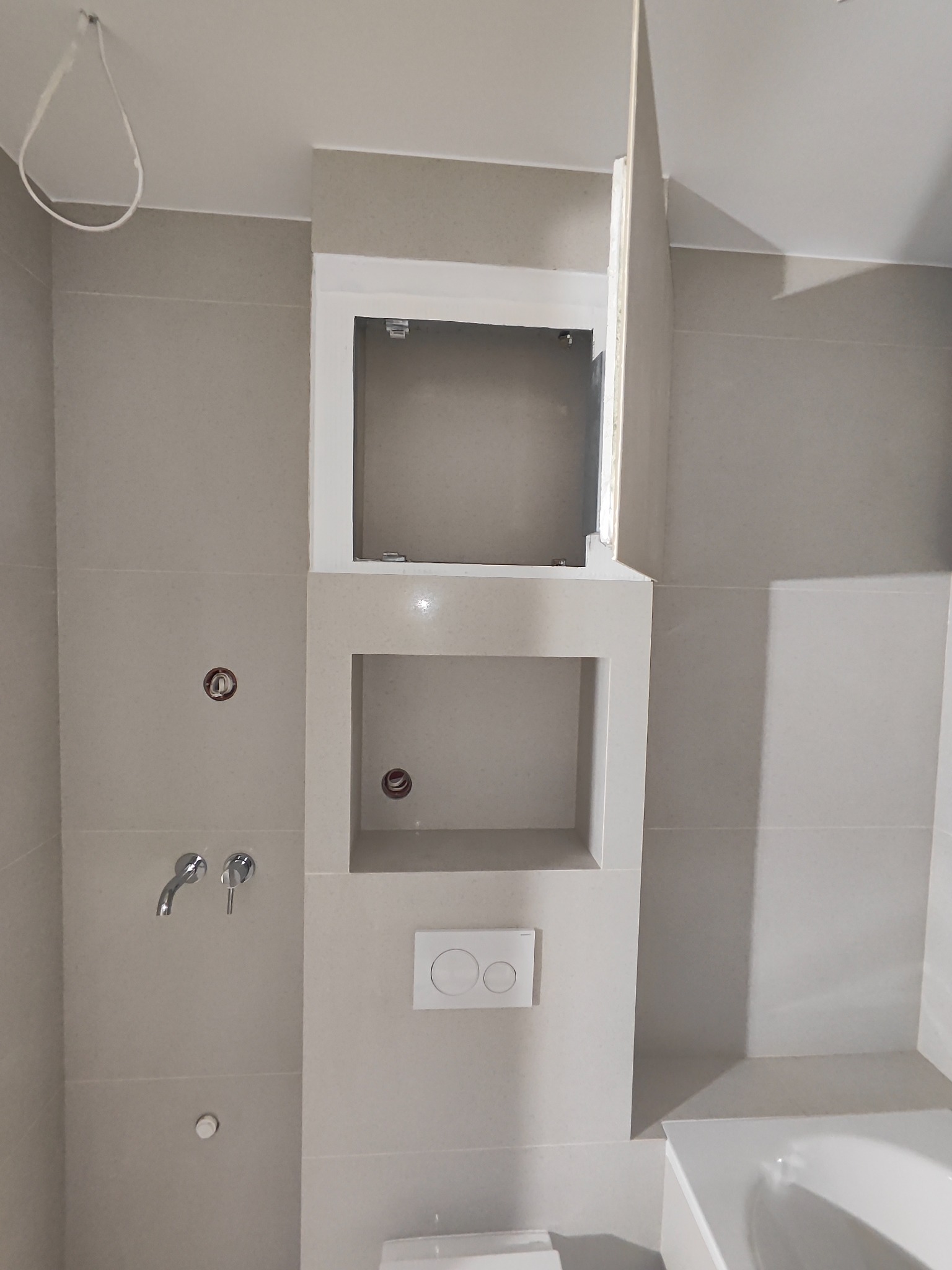 Wykończona łazienka z wnękami w ścianie wyłożonej płytkami w odcieniach szarości. Widoczna armatura, przycisk spłuczki i fragment wanny. Górna wnęka z otwartymi drzwiczkami.