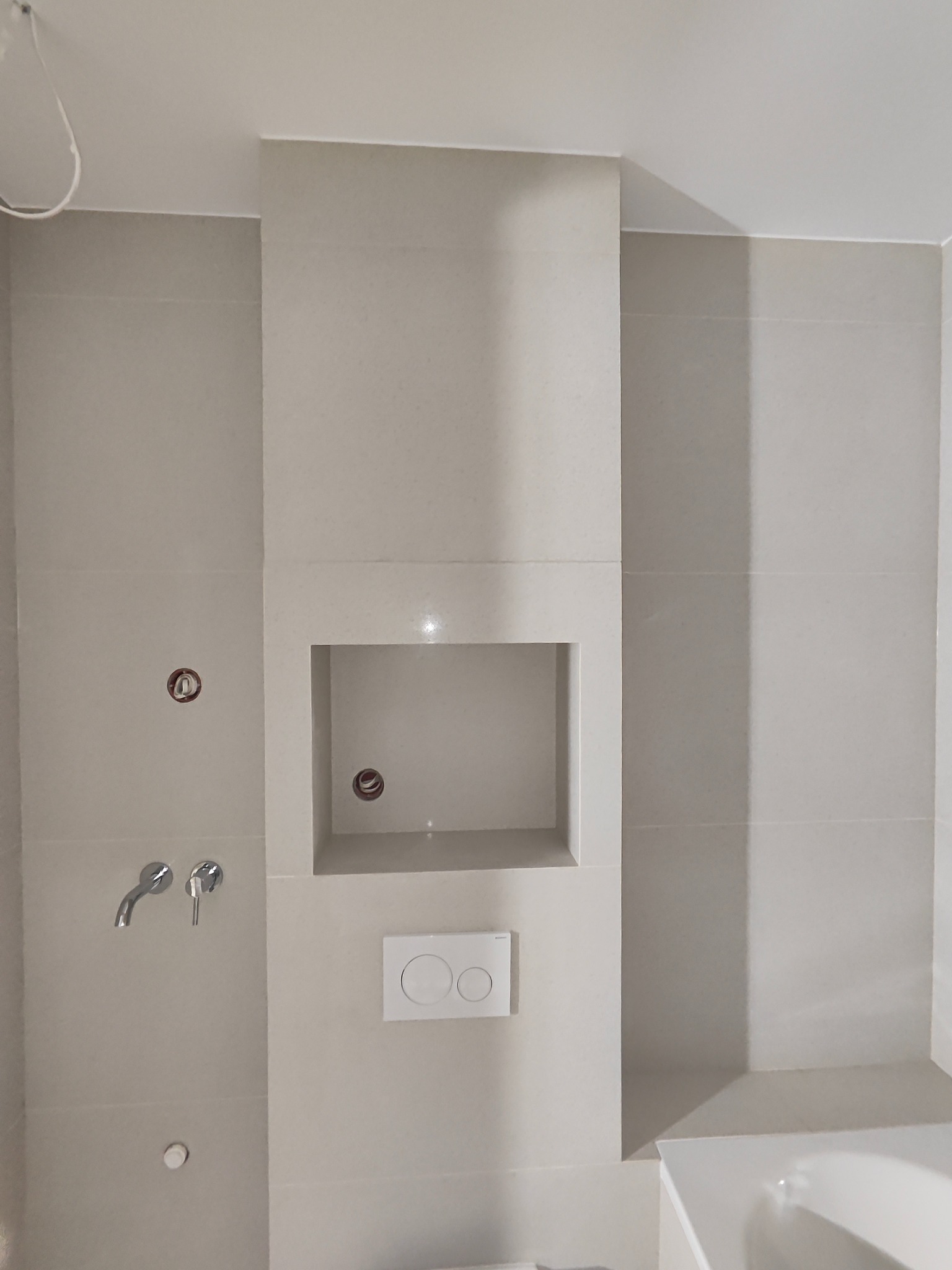 Wykończona łazienka z jasnymi płytkami, wnęką w ścianie i chromowaną armaturą. Widoczne przyłącza wodne i częściowo wanna. Minimalistyczny styl.