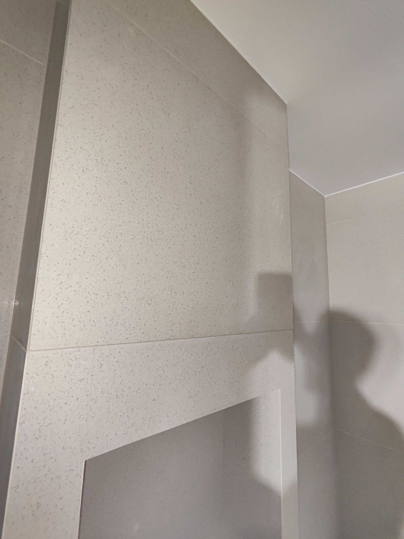 Wykończona ściana łazienki z jasnymi, wzorzystymi płytkami i wnęką. Minimalistyczny design, widoczny fragment sufitu. Detal wykończenia.