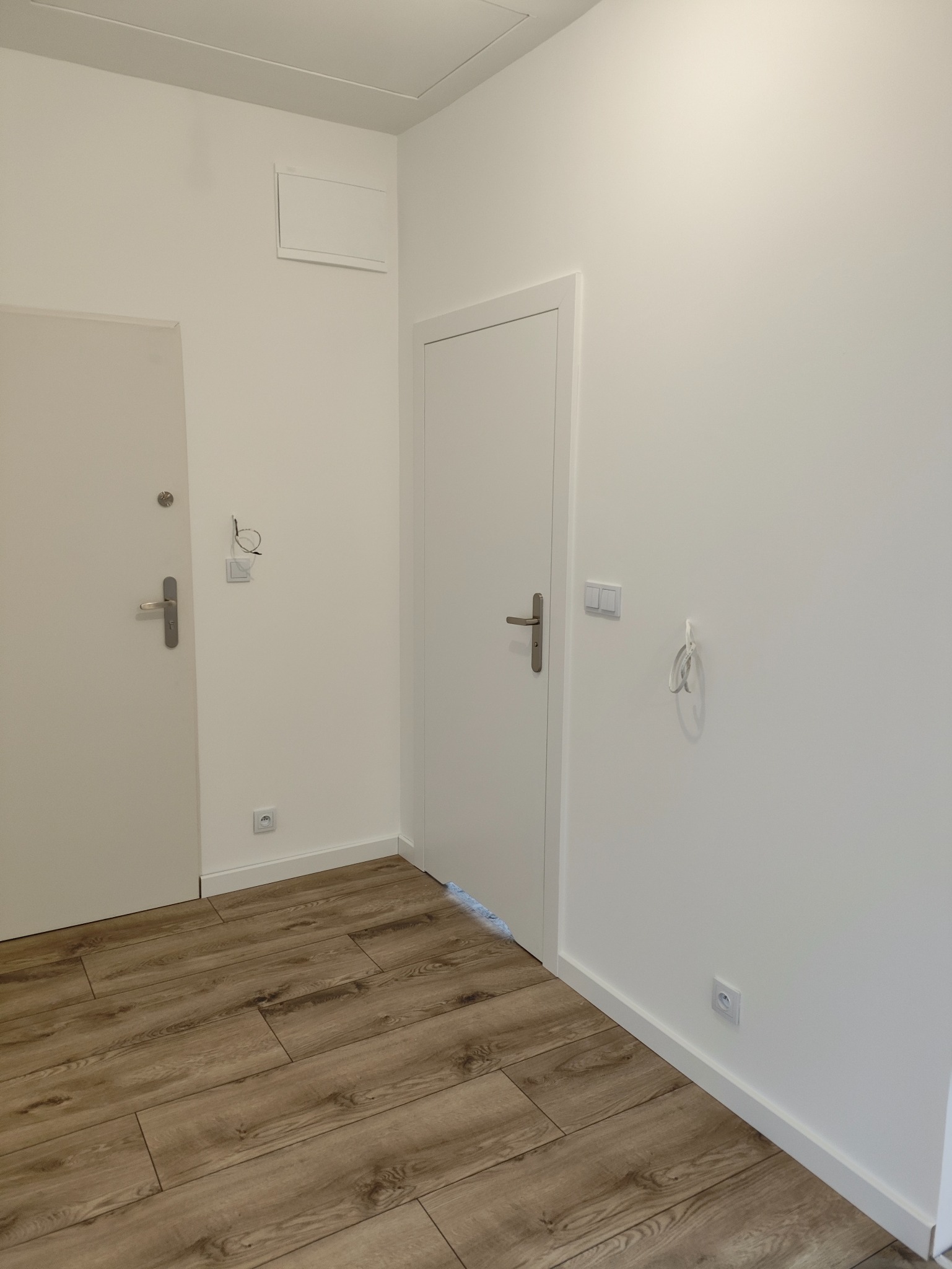 Białe wnętrze po remoncie: drewnopodobna podłoga, dwie pary drzwi, gładkie ściany z gniazdkami i włącznikami. Minimalistyczny design.