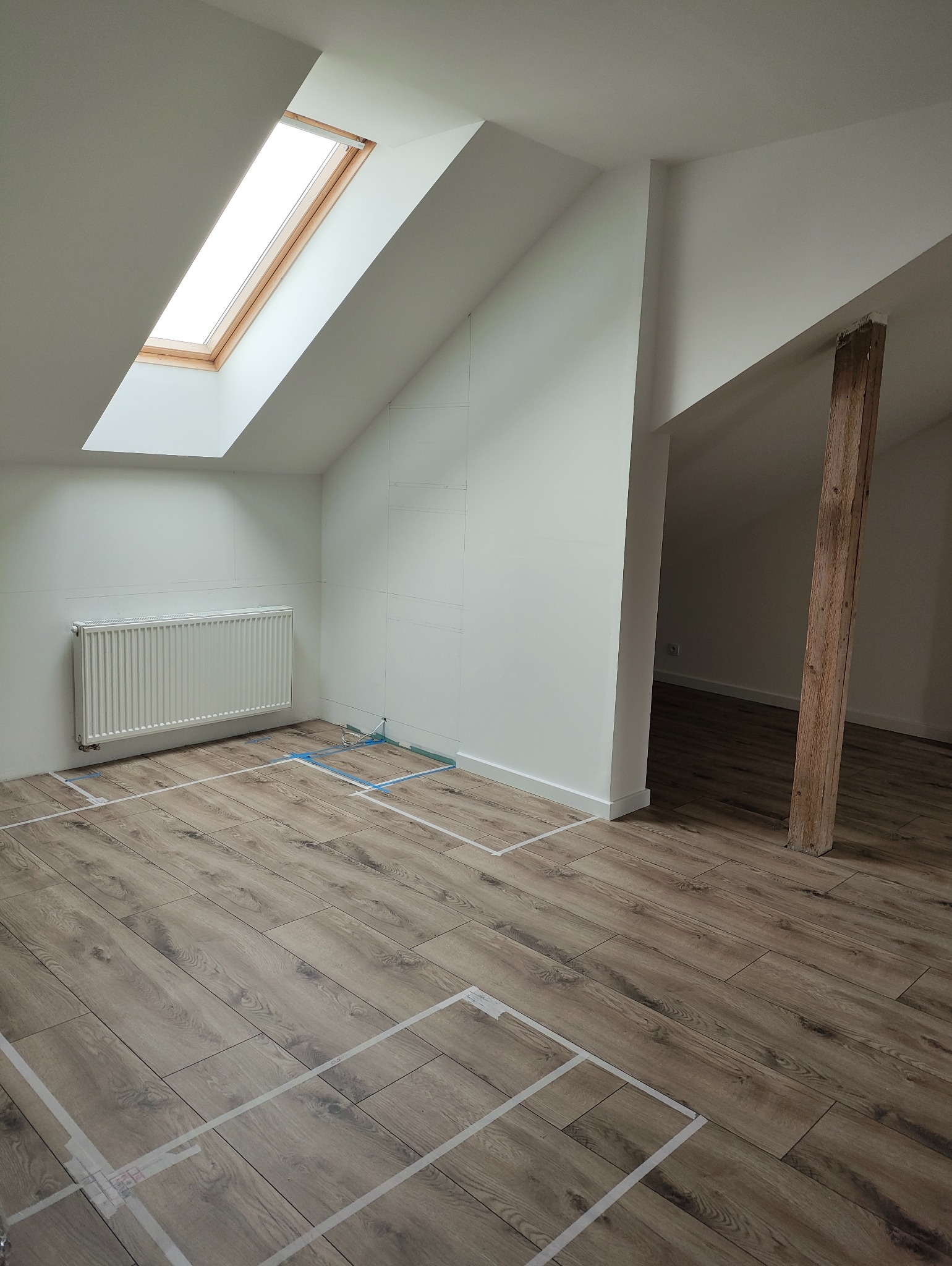 Podłoga z paneli ułożona w pokoju na poddaszu w Raszynie. Widoczne taśmy wyznaczające obszar roboczy, okno dachowe i grzejnik. Ściany przygotowane pod montaż płyt.