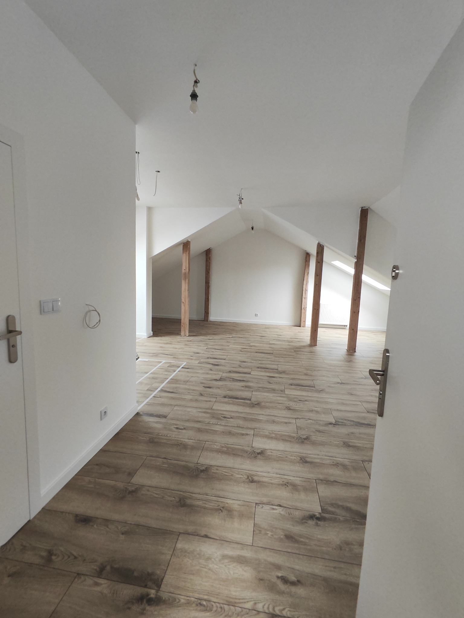 Wykończone poddasze z drewnopodobną podłogą i białymi ścianami. Widoczne drewniane słupy konstrukcyjne i niedokończone instalacje elektryczne.