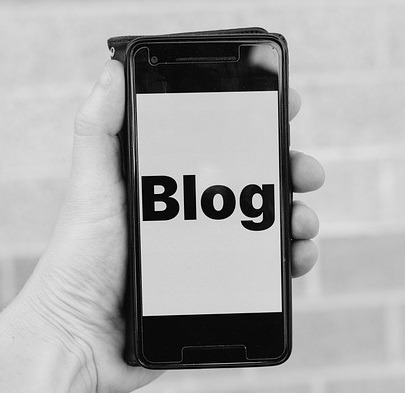 Czarno-białe zdjęcie dłoni trzymającej smartfon z wyświetlonym słowem 'Blog' na ekranie, na tle ceglanej ściany w Warszawie.