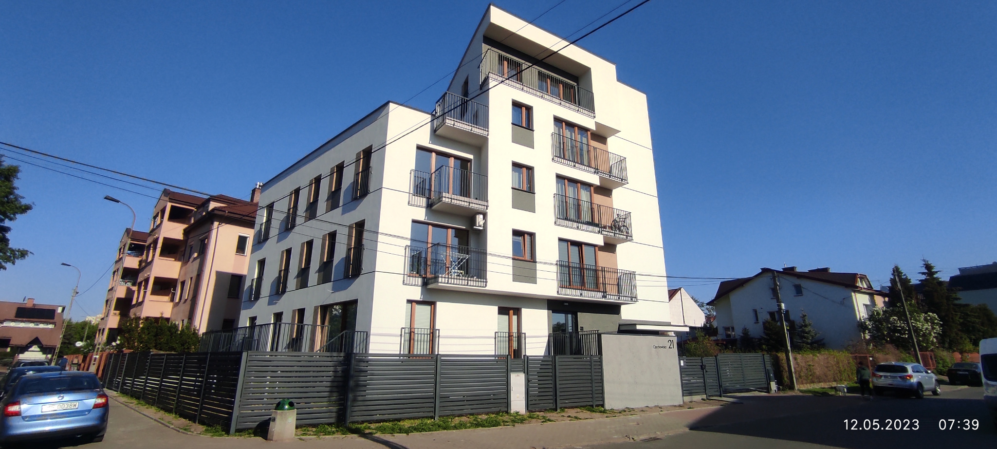 Nowoczesny, trzypiętrowy apartamentowiec z balkonami i szarym ogrodzeniem panelowym, sfotografowany w słoneczny poranek.