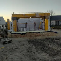 Wznoszenie ścian z bloczk&oacute;w betonu kom&oacute;rkowego; widoczne ż&oacute;łte szalunki i stalowe zbrojenie. W tle budynek z cegły i palety z bloczkami.