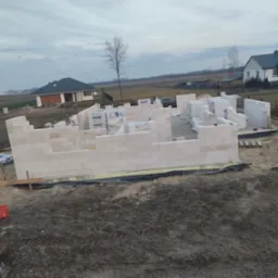 Wznoszenie ścian parterowego domu z bloczków Solbet. Na pierwszym planie fragment fundamentów. W tle widoczne inne budynki i pochmurne niebo.
