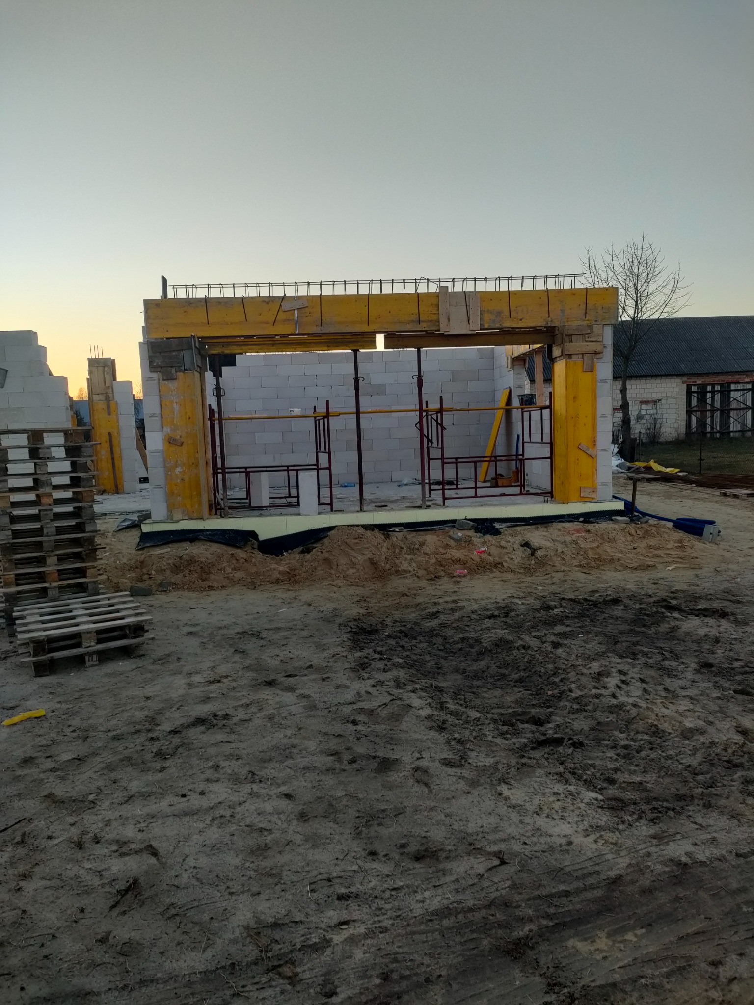 Wznoszenie ścian z bloczków betonu komórkowego; widoczne żółte szalunki i stalowe zbrojenie. W tle budynek z cegły i palety z bloczkami.