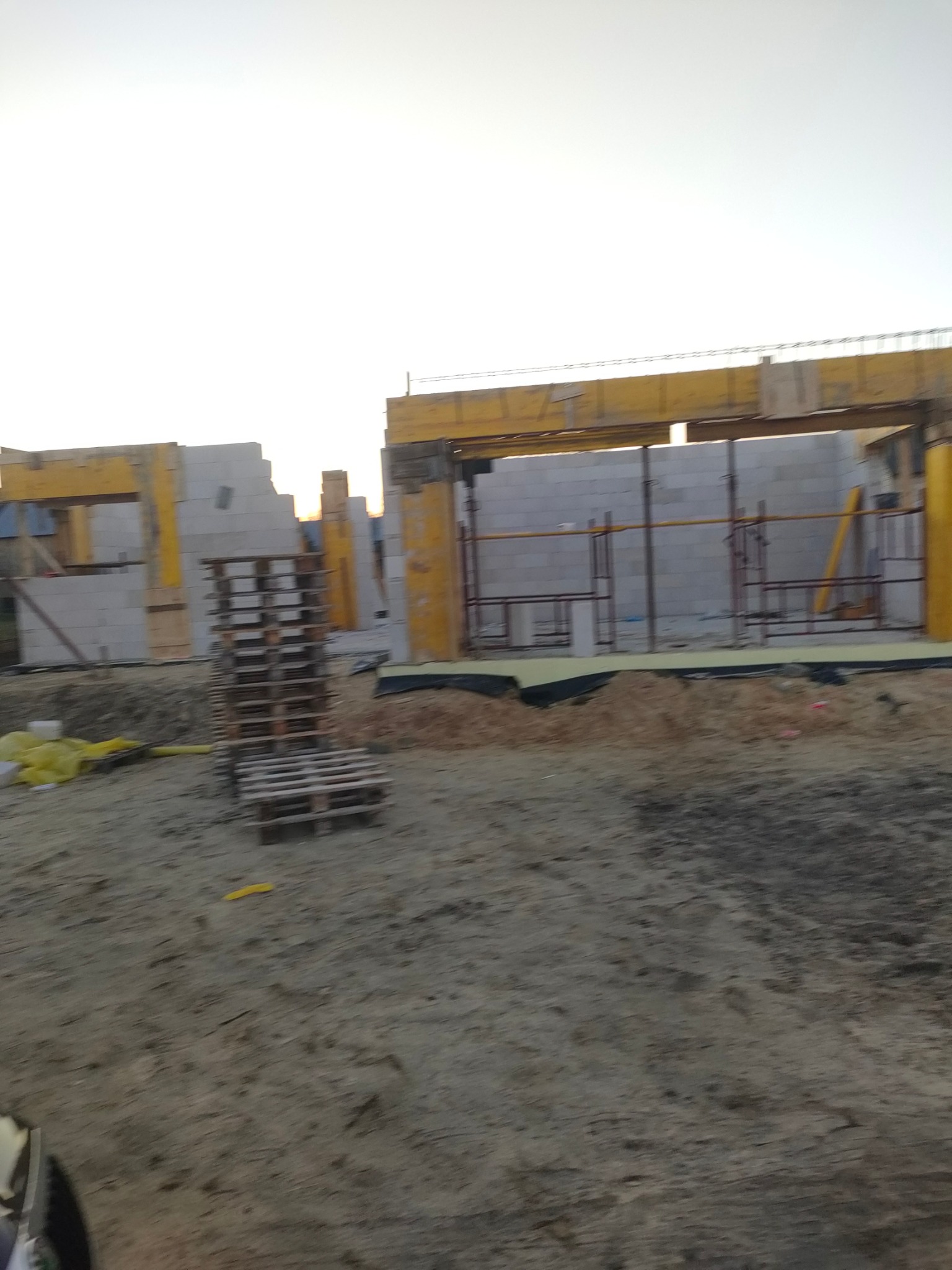 Budowa domu z bloczków: ściany w trakcie wznoszenia, widoczne żółte podpory i drewniane palety na placu budowy, w tle jasne niebo.