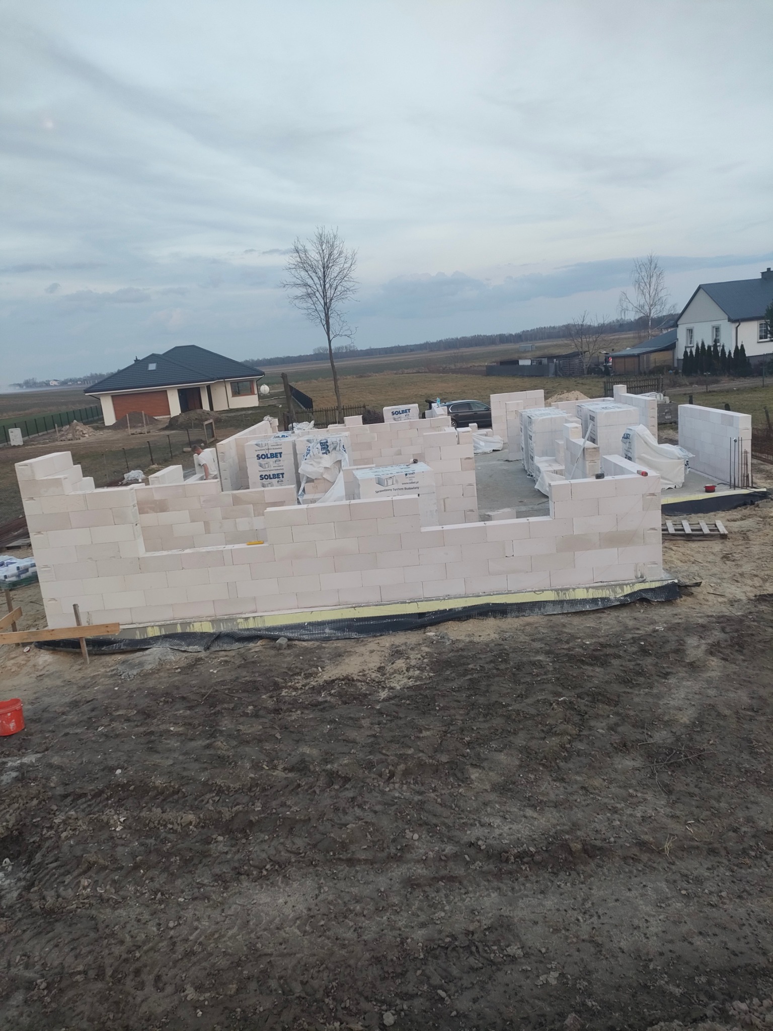 Wznoszenie ścian parterowego domu z bloczków Solbet. Na pierwszym planie fragment fundamentów. W tle widoczne inne budynki i pochmurne niebo.