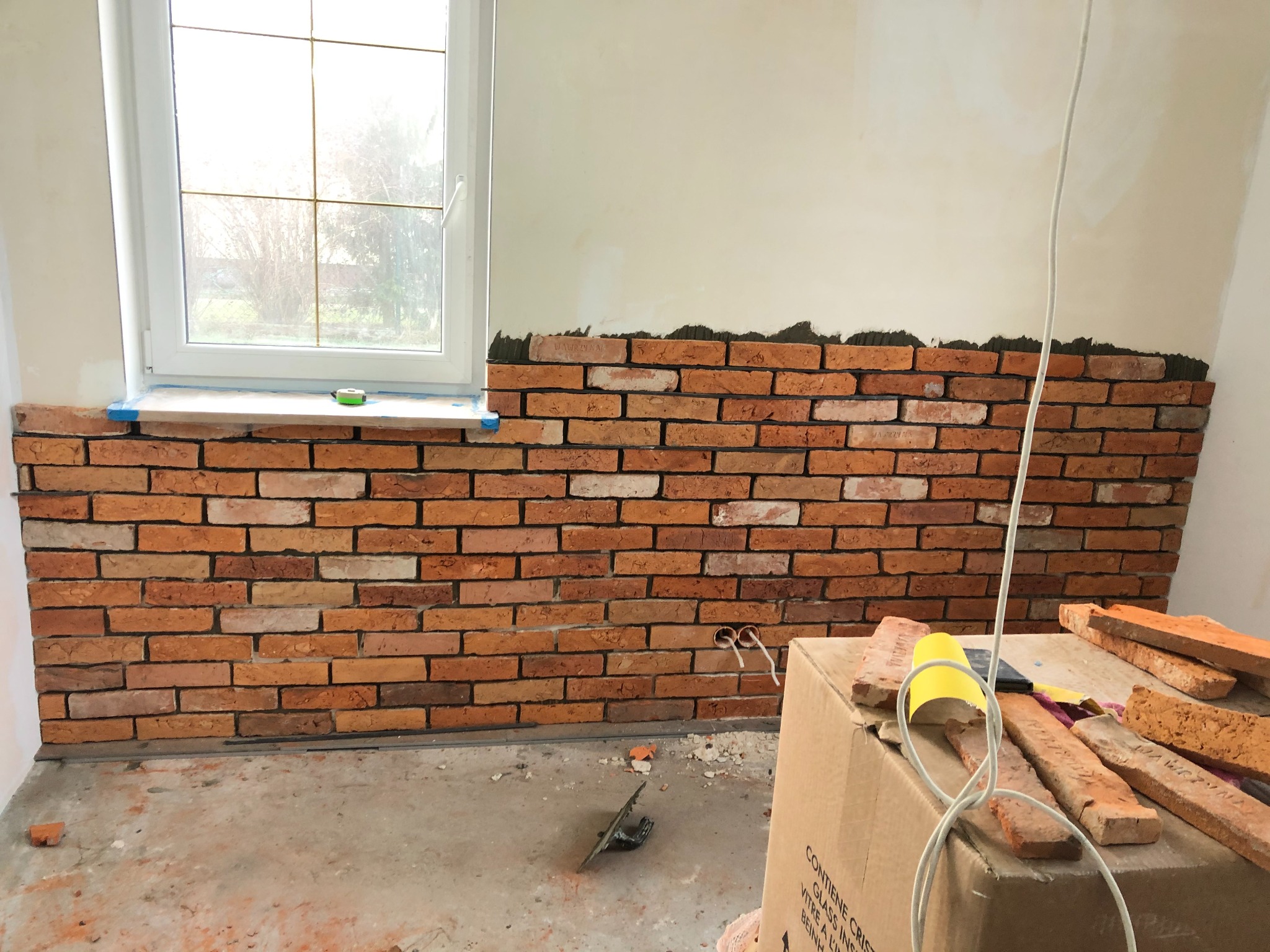 Montaż dekoracyjnej ściany z cegły w pomieszczeniu z oknem, widoczne narzędzia i materiały na kartonie, częściowo wykończona ściana z widocznym klejem.