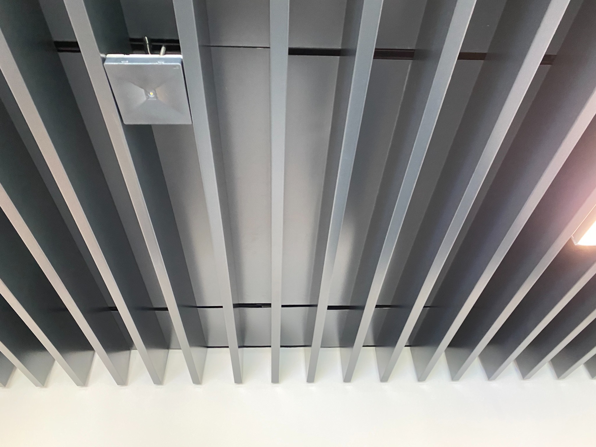 Szare, prostopadłe panele sufitowe tworzące geometryczny wzór, z widocznym kwadratowym reflektorem LED.