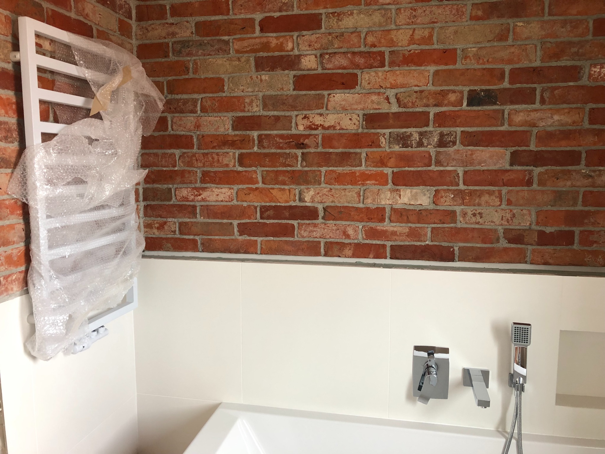 Nowoczesna łazienka w trakcie wykańczania: biała wanna, armatura w zabudowie, ściana z czerwonej cegły, białe płytki, grzejnik łazienkowy owinięty folią bąbelkową.