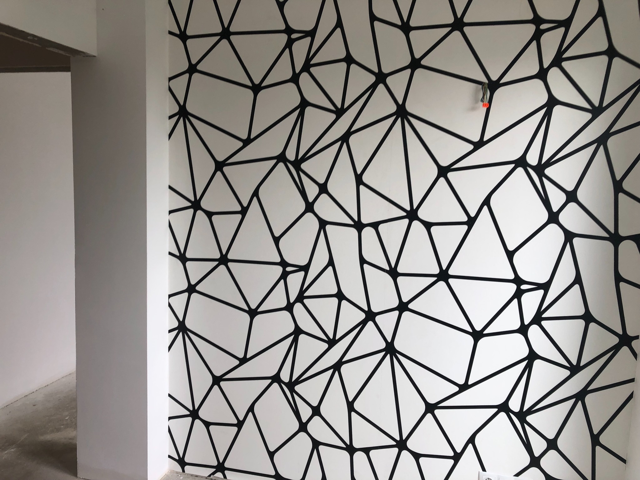 Biała ściana z czarnym geometrycznym wzorem przypominającym pajęczynę, widoczne wystające kable, surowy stan wykończenia wnętrza.