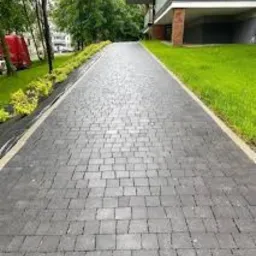 Długa, szara ścieżka ułożona z kostki granitowej prowadząca w kierunku nowoczesnego budynku z balkonem, otoczona zielenią i niskimi krzewami.